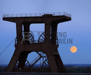 Das 55 Meter hohe Doppelbock-Fördergerüst ist das Wahrzeichen der Zeche Zollverein, der Stadt Essen und der gesamten Region: Die als Doppelstrebengerüst in Vollwandbauweise konstruierte Förderanlage ermöglichte eine doppelte Fördermechanik. Dadurch konnte die doppelte Menge an Förderwagen bzw. Fördergefäßen transportiert werden. Zollverein Schacht XII galt zu Betriebszeiten als die leistungsstärkste Zeche der Welt. 250 Stufen führen auf die oberste Plattform des Gerüstes, deren Seilscheiben sich auch heute noch manchmal drehen: Der Schacht wird von der RAG AG für die zentrale Wasserhaltung genutzt.

Areal A [Schacht XII], Fördergerüst [A1]